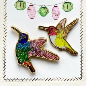 Könnte beinhalten: Zwei bunte Kolibri-Anstecker mit goldenen Akzenten. Der Kolibri links hat einen grünen, blauen und rosafarbenen Körper mit einem roten Schwanz. Der Kolibri rechts hat einen gelben Körper mit einem roten Kopf und braunen Flügeln.