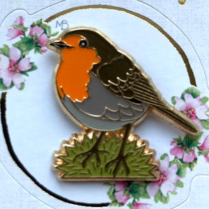 May include: A gold-colored robin pin with an orange breast, gray body, and brown wings. The bird is perched on a patch of green grass. The pin is set against a white background with floral accents.