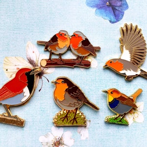 May include: Five enamel pins featuring different types of robins. The pins are all gold-toned and have a detailed design. The robins are depicted in various poses, including flying, perched, and singing.