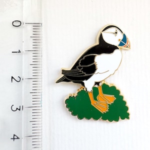 Könnte beinhalten: Ein farbenfroher Emaille-Pin mit einem Papageientaucher. Der Vogel hat einen schwarzen Rücken, einen weißen Bauch und einen markanten orangefarbenen Schnabel. Er sitzt auf einer grünen, blattreichen Basis mit goldenem Rand. Der Pin ist etwa 2,5 cm hoch.