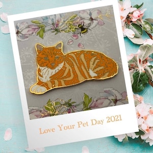Könnte beinhalten: Orangefarbene Tabby-Katzen-Enamel-Anstecknadel mit goldenen Akzenten. Die Anstecknadel liegt mit ausgestreckten Pfoten. Die Anstecknadel befindet sich auf einem floralen Hintergrund mit dem Text "Love Your Pet Day 2021".