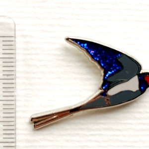 K&ouml;nnte beinhalten: Eine blaue und silberne Emaille-Anstecknadel mit einer fliegenden Schwalbe. Die Fl&uuml;gel der Schwalbe sind in einem glitzernden Blau gehalten, und ihr K&ouml;rper ist grau mit einem wei&szlig;en Bauch. Der Schnabel der Schwalbe ist rot.