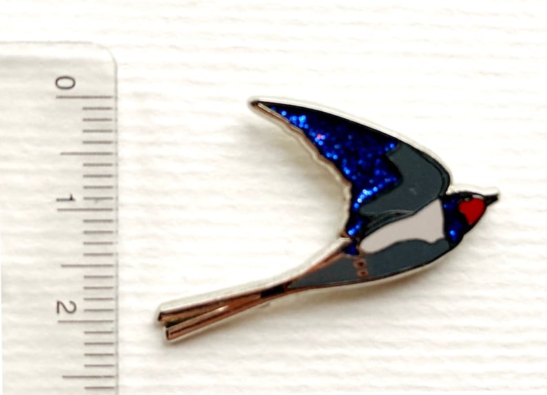 K&ouml;nnte beinhalten: Eine blaue und silberne Emaille-Anstecknadel mit einer fliegenden Schwalbe. Die Fl&uuml;gel der Schwalbe sind in einem glitzernden Blau gehalten, und ihr K&ouml;rper ist grau mit einem wei&szlig;en Bauch. Der Schnabel der Schwalbe ist rot.