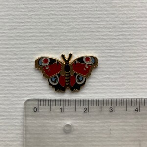 May include: A gold-colored metal pin of a red, black, and blue butterfly with white spots. The butterfly is about 1.5 inches wide.