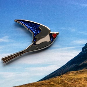 K&ouml;nnte beinhalten: Eine silberfarbene Emaille-Anstecknadel mit einem blauen und wei&szlig;en Vogel mit einem roten Herzen auf der Brust. Der Vogel ist im Flug mit ausgebreiteten Fl&uuml;geln. Die Anstecknadel ist vor einem blauen Himmel und einem Berg im Hintergrund platziert.