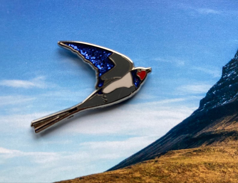 K&ouml;nnte beinhalten: Eine silberfarbene Emaille-Anstecknadel mit einem blauen und wei&szlig;en Vogel mit einem roten Herzen auf der Brust. Der Vogel ist im Flug mit ausgebreiteten Fl&uuml;geln. Die Anstecknadel ist vor einem blauen Himmel und einem Berg im Hintergrund platziert.