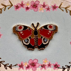 May include: Gold-tone metal pin featuring a red, black, and white butterfly with intricate details. The butterfly is set against a light blue background with a floral border.