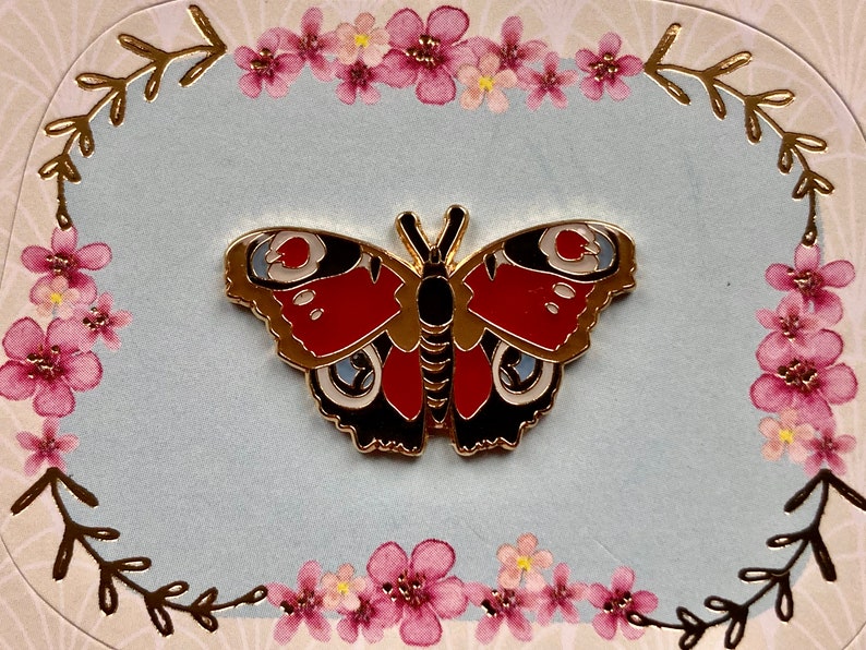 May include: Gold-tone metal pin featuring a red, black, and white butterfly with intricate details. The butterfly is set against a light blue background with a floral border.