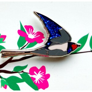 K&ouml;nnte beinhalten: Eine silberne Emaille-Anstecknadel mit einem blauen und grauen Vogel mit roten und wei&szlig;en Akzenten. Der Vogel fliegt mit ausgebreiteten Fl&uuml;geln vor einem Hintergrund aus rosa Bl&uuml;ten und gr&uuml;nen Bl&auml;ttern.