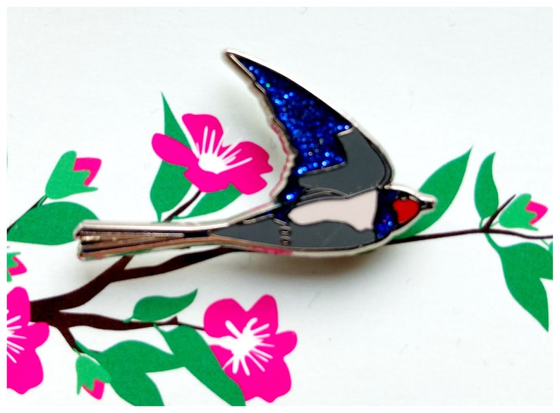 K&ouml;nnte beinhalten: Eine silberne Emaille-Anstecknadel mit einem blauen und grauen Vogel mit roten und wei&szlig;en Akzenten. Der Vogel fliegt mit ausgebreiteten Fl&uuml;geln vor einem Hintergrund aus rosa Bl&uuml;ten und gr&uuml;nen Bl&auml;ttern.