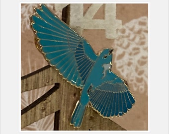 May include: A blue enamel pin of a bird with its wings spread. The bird is in flight and has a white belly. The pin is on a wooden background.