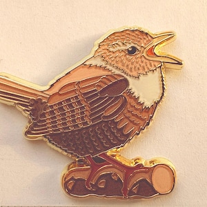 Könnte beinhalten: Ein detaillierter, goldfarbener Vogel-Ansteckpin mit braunen und beigen Federn, der auf einem braunen Baumstamm sitzt. Der Vogel hat einen offenen Schnabel mit roter Innenseite, und der Pin hat eine polierte Oberfläche. Das Design ist aufwendig und auffällig.