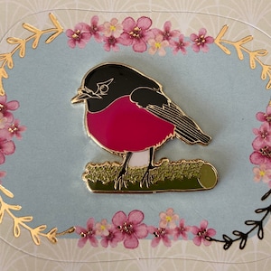 Könnte beinhalten: Eine goldfarbene Emaille-Anstecknadel mit einem Rotkehlchen, das auf einem grünen Zweig sitzt. Das Rotkehlchen hat einen schwarzen Kopf, eine rosafarbene Brust und einen weißen Bauch. Die Anstecknadel ist von einem floralen Rand umgeben.