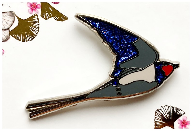 K&ouml;nnte beinhalten: Eine silberne Emaille-Anstecknadel mit einer blauen und grauen Schwalbe mit einem roten Herzen auf der Brust. Die Fl&uuml;gel der Schwalbe sind mit blauen Glitzer bedeckt.