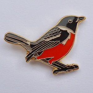 May include: A gold-colored metal pin of a black and white bird with an orange breast. The bird is perched on a branch with its wings spread.