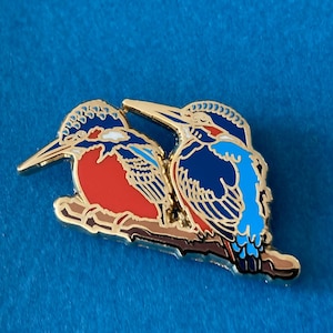 Op de afbeelding: Een gouden emaille pin met twee ijsvogels die op een tak zitten. De vogels kijken elkaar aan, een in blauw en turquoise en de andere in rood en blauw.