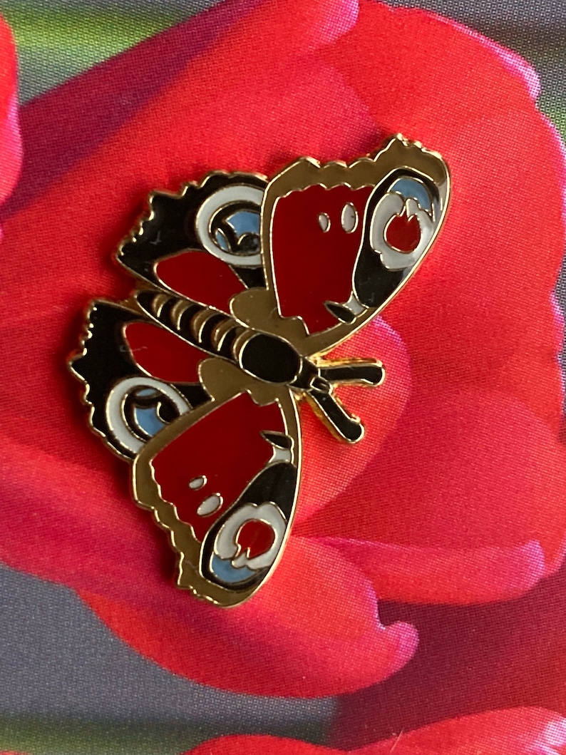 May include: A gold-colored metal pin with a red, black, and blue butterfly design. The butterfly has white spots on its wings.