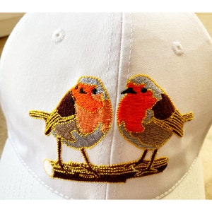 Puede incluir: Gorra de béisbol blanca con petirrojos bordados posados en una rama.