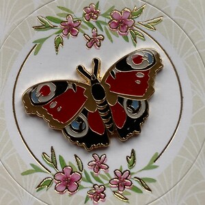 May include: A gold-colored butterfly pin with red, black, and white details. The butterfly is surrounded by a floral design with pink flowers and green leaves.