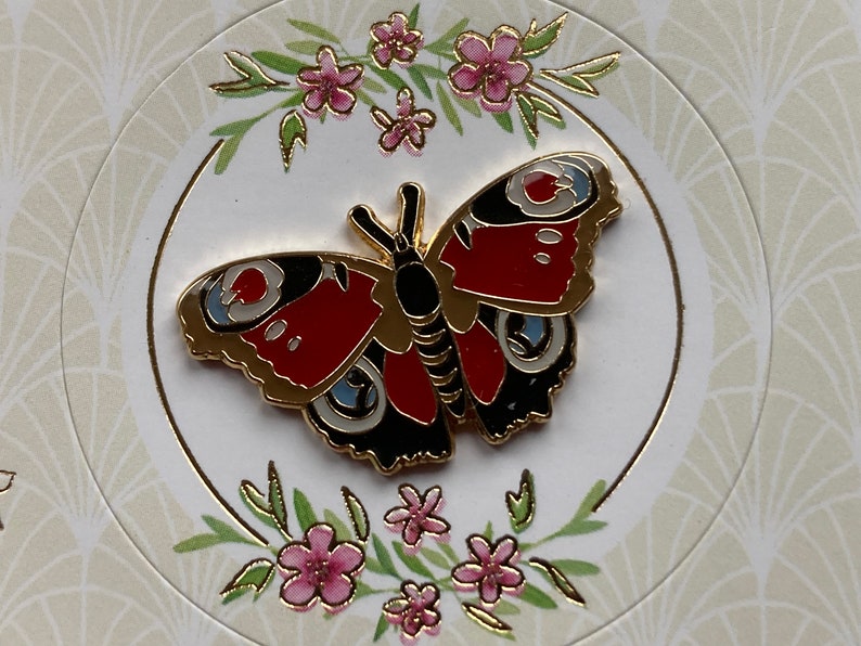 May include: A gold-colored butterfly pin with red, black, and white details. The butterfly is surrounded by a floral design with pink flowers and green leaves.