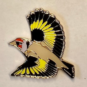 Op de afbeelding: Een gouden metalen pin van een gele en zwarte vogel met een rood-witte kop in vlucht.