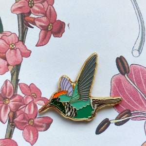 Op de afbeelding: Een gouden emaille pin met een kolibrie in vlucht. De kolibrie heeft groene, paarse en oranje accenten.