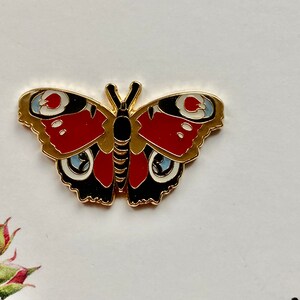 May include: Gold-tone metal pin featuring a red, black, and white butterfly with a detailed design.