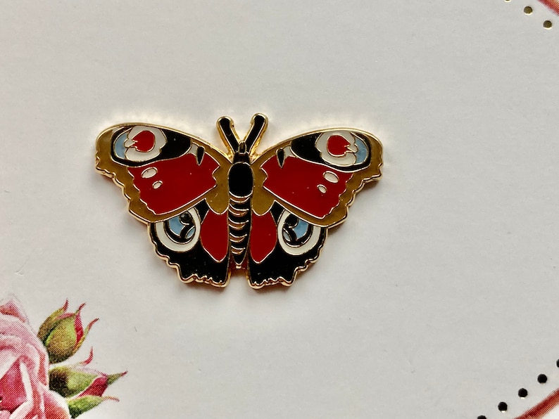 May include: Gold-tone metal pin featuring a red, black, and white butterfly with a detailed design.