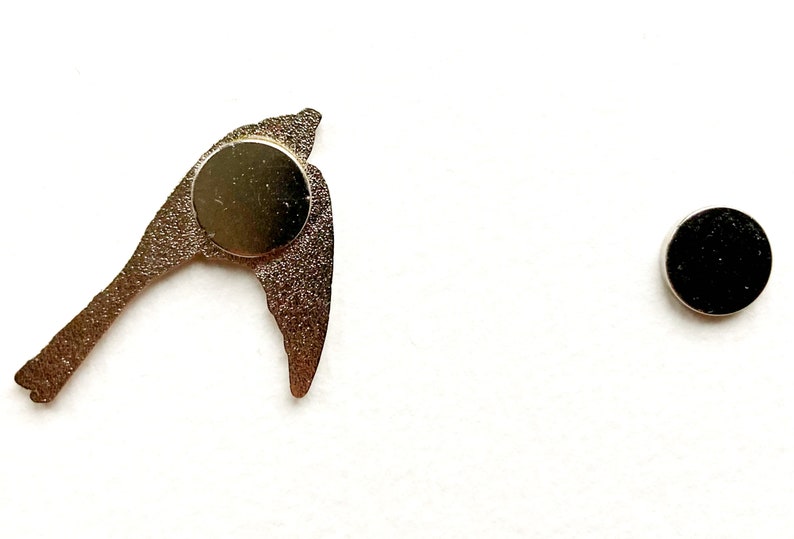 K&ouml;nnte beinhalten: Eine goldfarbene Metall-Vogelsilhouette mit einem runden Magneten auf der R&uuml;ckseite. Der Magnet ist schwarz und hat eine strukturierte Oberfl&auml;che.