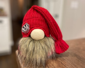 Puede incluir: Un gorro de gnomo de punto rojo con un parche de logotipo de tejón blanco y negro. El gnomo tiene una larga barba de piel marrón y negra.