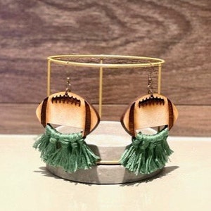 May include: A pair of wooden football earrings with green tassel fringe. The earrings are displayed on a gold stand.