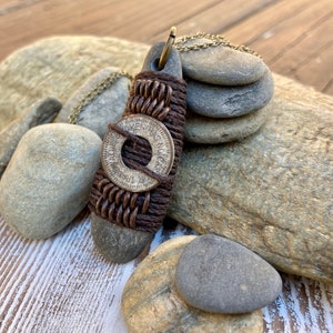 May include: A brown leather cord necklace with a silver coin pendant. The coin has the words "Token Has No Cash Value" inscribed on it. The necklace is displayed on a wooden surface with several gray stones.