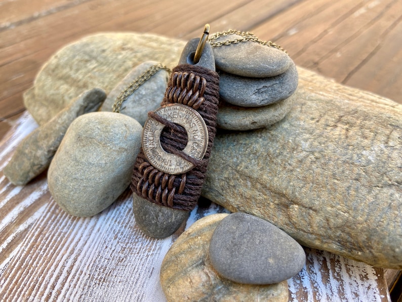 May include: A brown leather cord necklace with a silver coin pendant. The coin has the words "Token Has No Cash Value" inscribed on it. The necklace is displayed on a wooden surface with several gray stones.