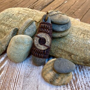 May include: A brown leather and metal necklace with a round pendant featuring the text "Token Has No Value". The necklace is displayed on a wooden surface with rocks.