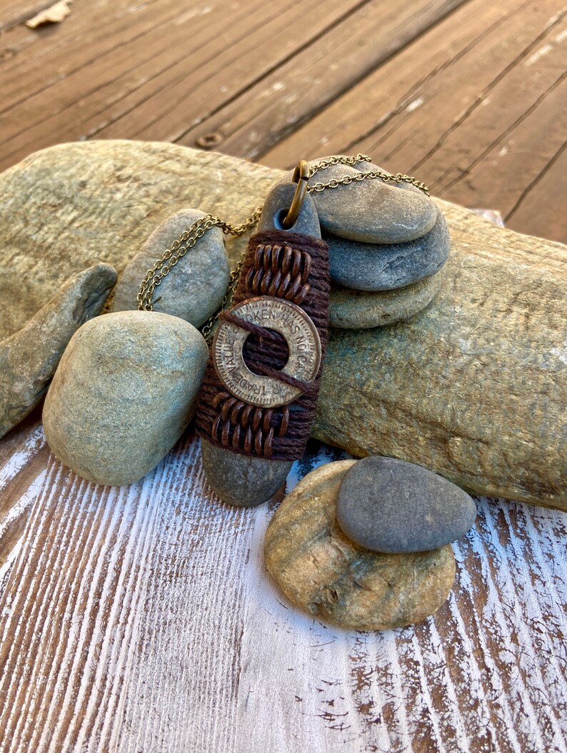 May include: A brown leather and metal necklace with a round pendant featuring the text "Token Has No Value". The necklace is displayed on a wooden surface with rocks.