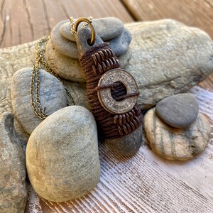 May include: A brown leather cord necklace with a stone pendant wrapped in brown leather cord and a silver token with the text "Token Has No Value" and a circle in the center. The necklace is displayed on a pile of gray stones.