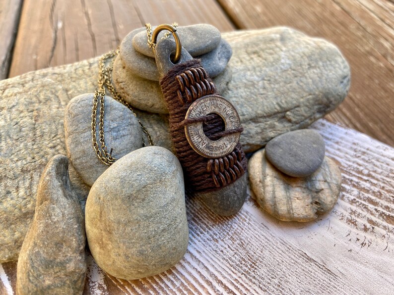 May include: A brown leather cord necklace with a stone pendant wrapped in brown leather cord and a silver token with the text "Token Has No Value" and a circle in the center. The necklace is displayed on a pile of gray stones.