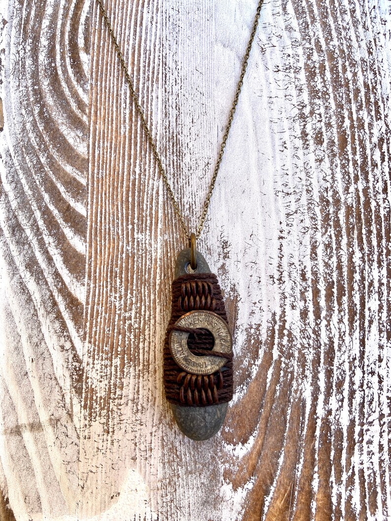 May include: A brown stone pendant necklace with a brass coin wrapped in brown leather cord. The coin has the words "Trade Token" inscribed on it.
