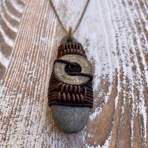 May include: A brown leather cord necklace with a gray stone pendant. The stone is wrapped with brown cord and a silver coin with the text "TRADE DOLLARS" is centered on the stone.