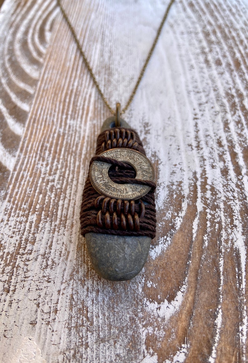 May include: A brown leather cord necklace with a gray stone pendant. The stone is wrapped with brown cord and a silver coin with the text "TRADE DOLLARS" is centered on the stone.