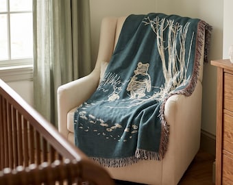 May include: A dark teal woven throw blanket featuring a cream-coloured line art illustration of Winnie the Pooh and Piglet standing near trees. The blanket has a fringed edge and is draped over a cream-coloured upholstered armchair.