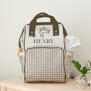 May include: A beige and brown diaper bag with a gingham pattern and a brown handle. The bag features a fish graphic and the name "HENRY" in capital letters. A small white hat is attached to the side.