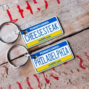 May include: Two rectangular metal keychains designed like Pennsylvania number plates. One reads CHEESESTEAK and the other PHILADELPHIA, both featuring the text PENNSYLVANIA and visitPA.com in blue, white, and yellow colours with silver key rings.