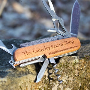 Könnte beinhalten: Ein Multifunktions-Taschenmesser mit einem Holzgriff, der mit "The Laundry Room Shop" und der Website-Adresse "info@thelaundryroomshop.com" graviert ist.