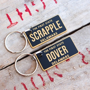 May include: Two keychains with rectangular black tags and gold lettering. One reads "SCRAPPLE DELAWARE" and the other "DOVER DELAWARE". Both say "THE FIRST STATE". The keychains are on a weathered wooden surface.