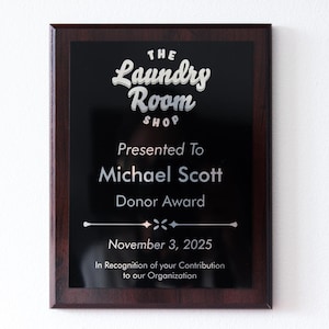 May include: A framed plaque with a dark background and a wooden frame. The text "The Laundry Room Shop" is at the top, followed by "Presented To Michael Scott Donor Award." The date "November 3, 2025" is also displayed.