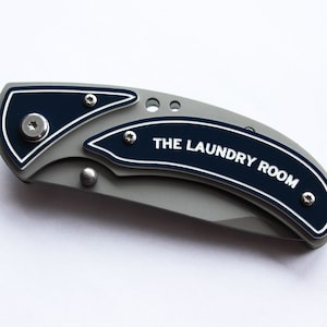 Puede incluir: Un cuchillo de bolsillo gris con mango azul. El mango tiene una inscripción blanca que dice "THE LAUNDRY ROOM".