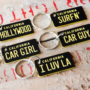 Puede incluir: Cinco llaveros negros y amarillos con diferentes frases con temática de California. Los llaveros presentan las frases "California Hollywood", "California Surfn'", "California Car Guy", "California Car Girl" y "California I Luv LA".