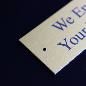 K&ouml;nnte beinhalten: Goldfarbene Metallplakette mit einem Loch in der oberen rechten Ecke. Die Plakette tr&auml;gt den Text "We Engrave Your Text" in blauer Schrift.