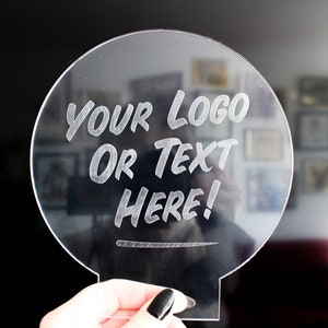 May include: Clear acrylic sign with a circular top and a rectangular base. The sign features the engraved text "YOUR LOGO OR TEXT HERE!". The background is blurred, with a dark area on the left and a wall with framed pictures on the right.